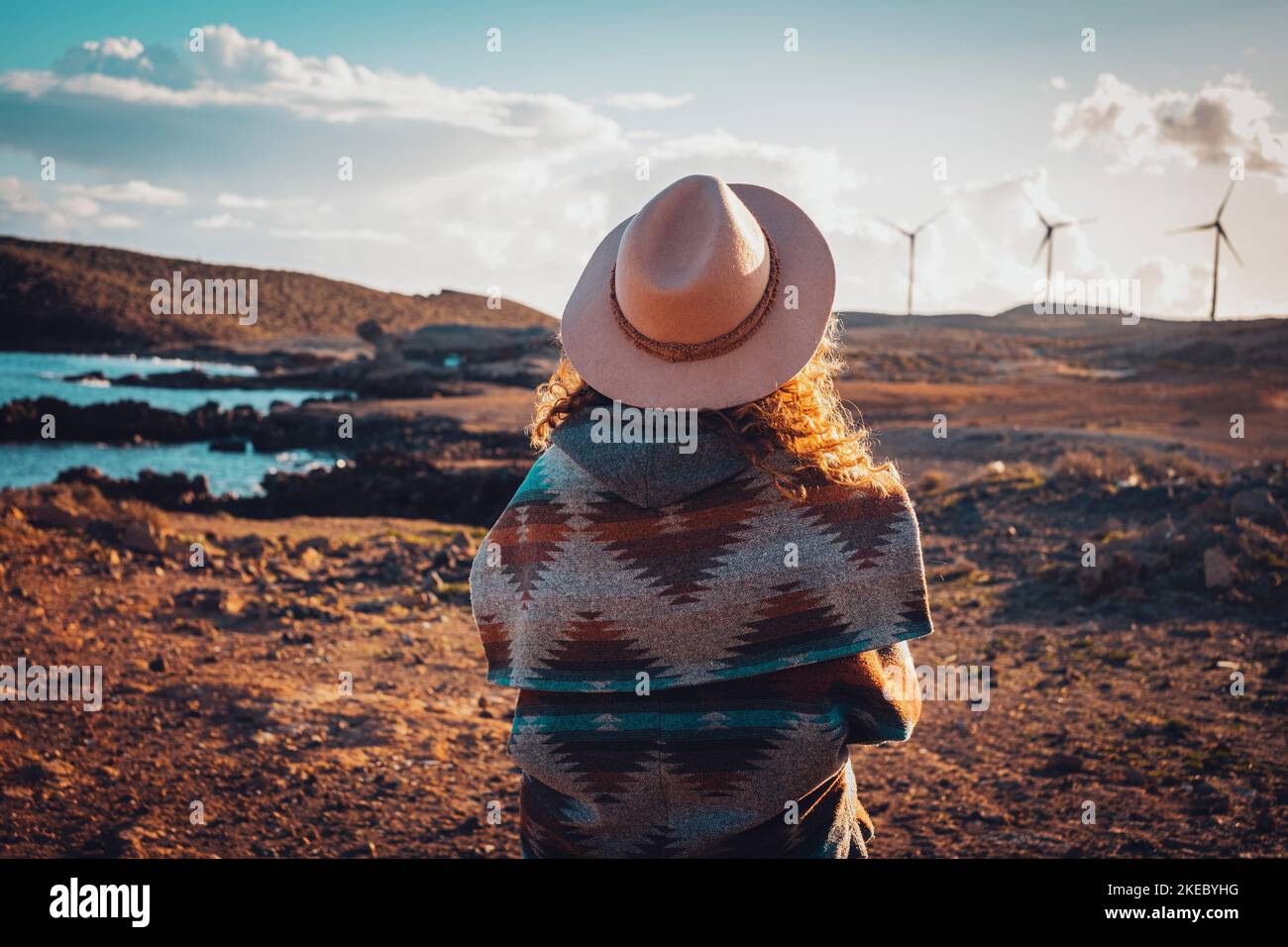 Back view of woman admiring landscape with windmills turbines in background. Concept of travel and wanderlust people in scenic place during sunset time. Summer vacation trip female. Blue sky and sea Stock Photo