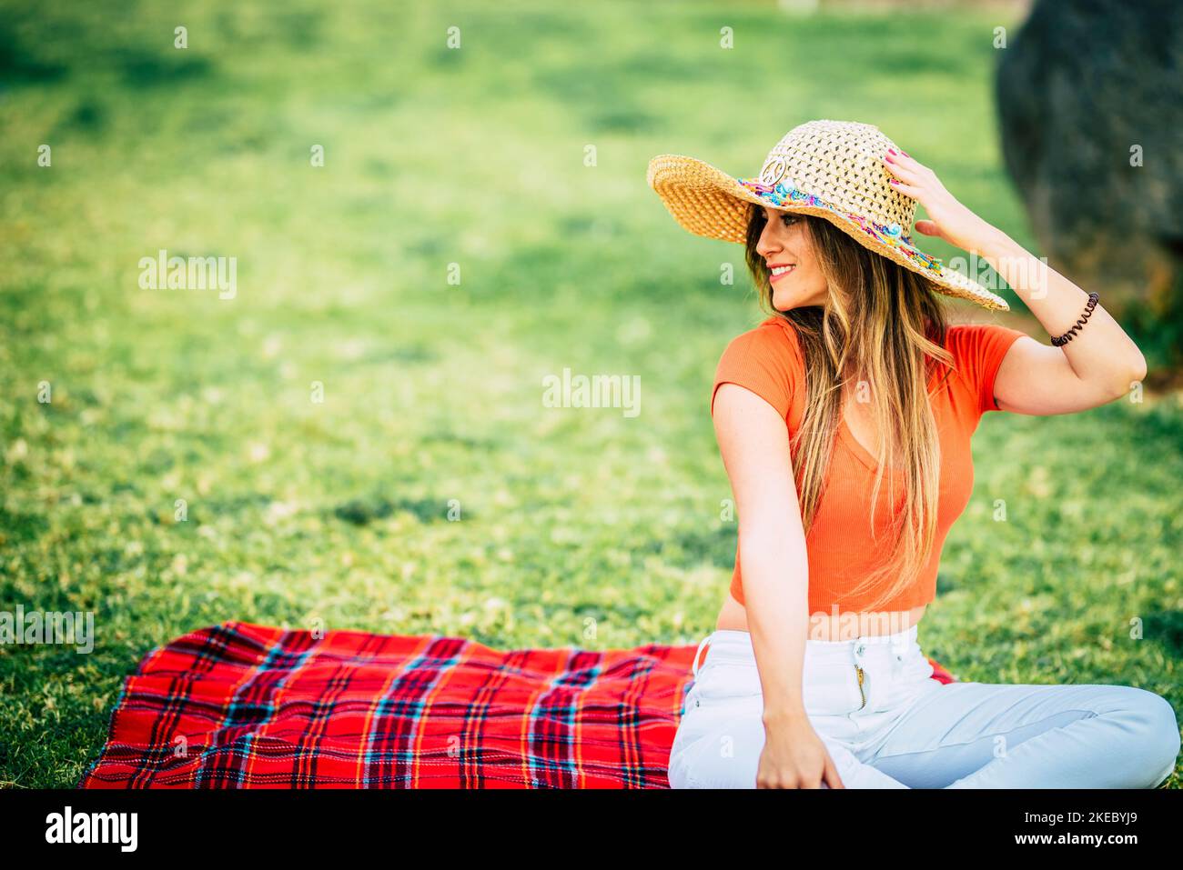 Portrait of trendy young female relaxing sitting on a red cover in a meadow field wearing straw hat enjoying time and the green nature. Concept of alone leisure serene outdoors activity at the park Stock Photo