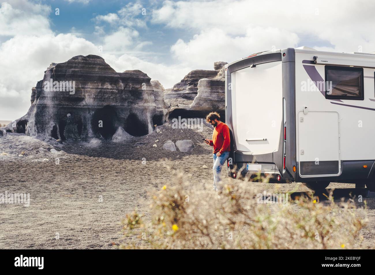 Standing man using mobile phone against a modern camper van and country side rural landscape in background. Concept of travel and connection technlogy to plan road trip and holiday vacation Stock Photo