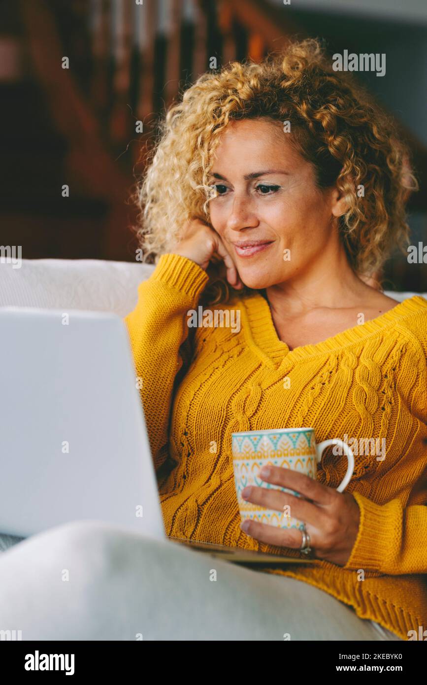 Female sitting with a cip of coffee on a couch, using laptop and internet connection and smile. Stock Photo