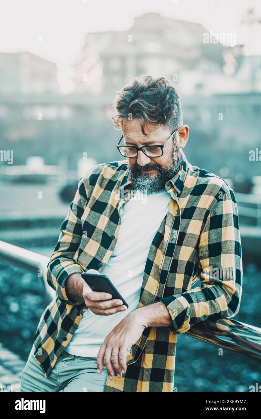 Mature man write and read on mobile phone standing outdoor. Technology and modern people concept lifestyle. Hipster style with beard male and glasses use app on cellular and send message chat Stock Photo