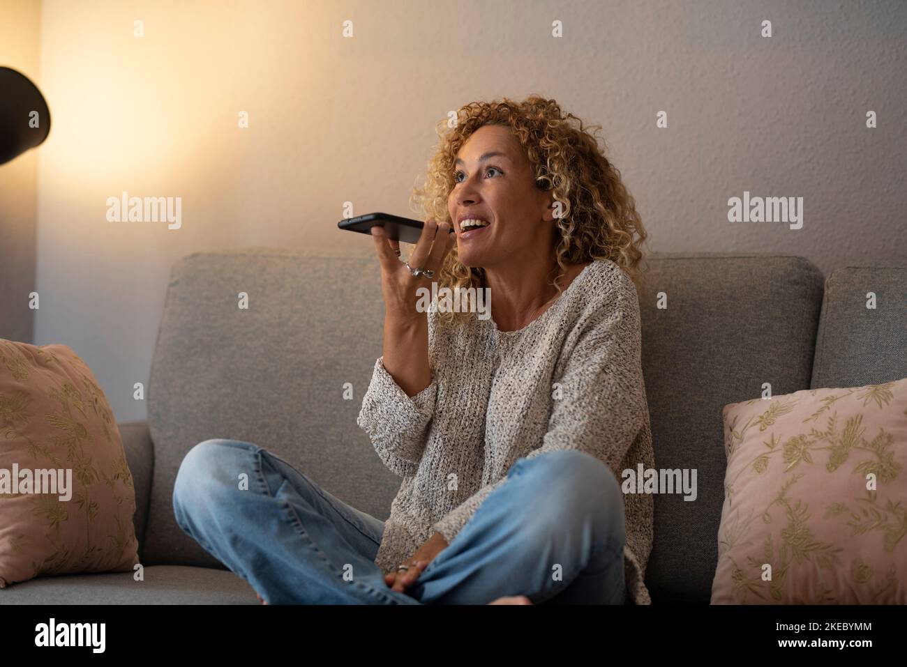 Excited woman speaking at the phone during cellular call at home sitting on the sofa. Indoor leisure activity and use of modern device. Female people enjoying voice connection with friends. Smartphone Stock Photo