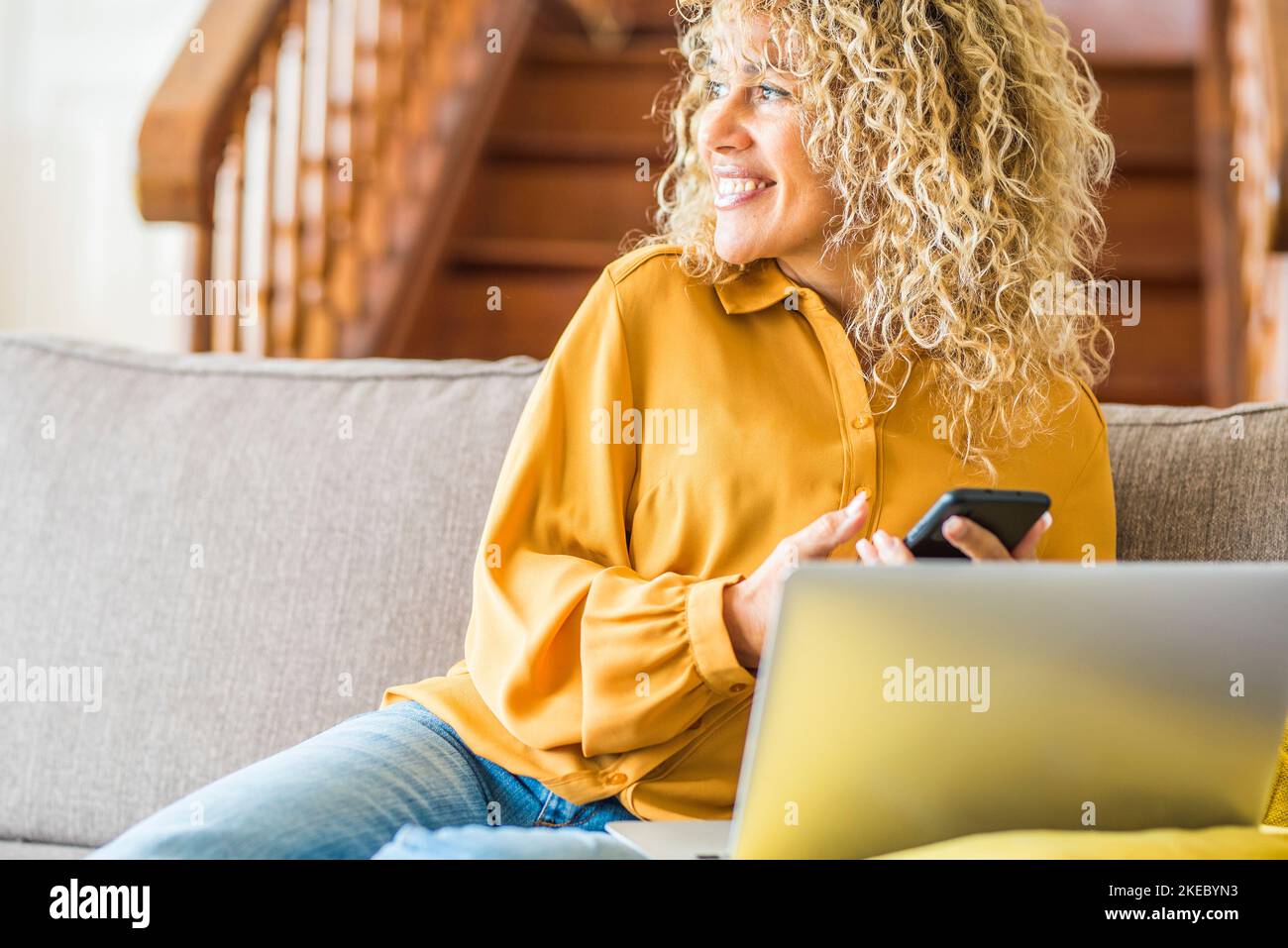 Happy attractive woman smiling and using mobile phone and laptop on the sofa at home. Adult young female smile and look on her side. Indoor technology leisure activity with cellular and computer Stock Photo