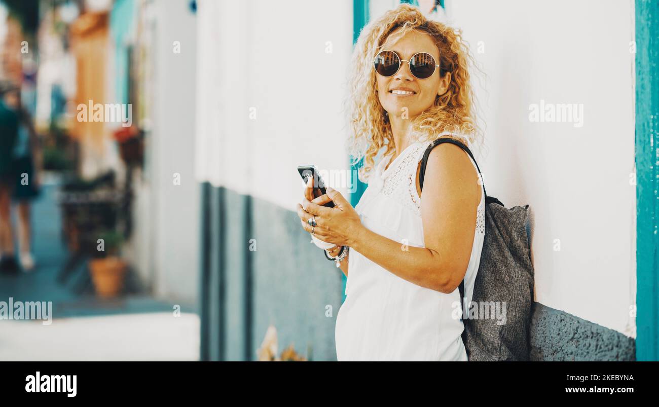 Happy female tourist pose for a picture using mobile phone connection against a white wall. Side portrait of woman smiling and writing on the cellular. Outdoor town leisure activity lady in summer Stock Photo