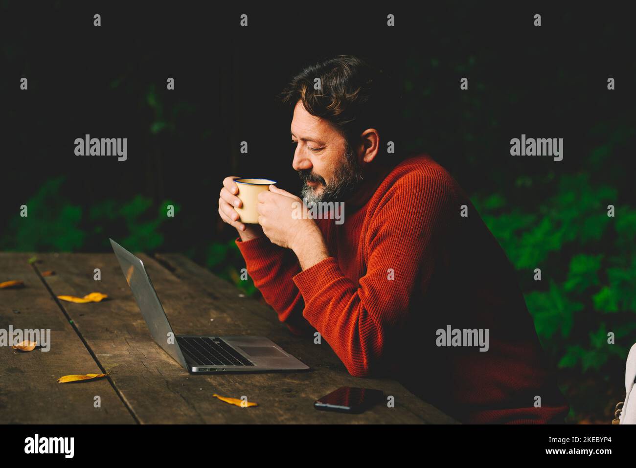 One man using laptop outdoor at the park sitting on a wooden bench with green woods background. Internet connection everywhere and digital nomad job lifestyle people. Using computer and phone device Stock Photo