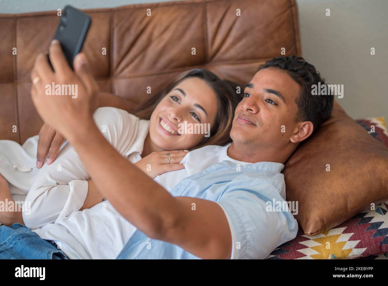 Young couple in love enjoy and use modern mobile phone together laying and relaxing on the sofa. Love and relationship young people indoor leisure activity with internet connection. Video call concept Stock Photo