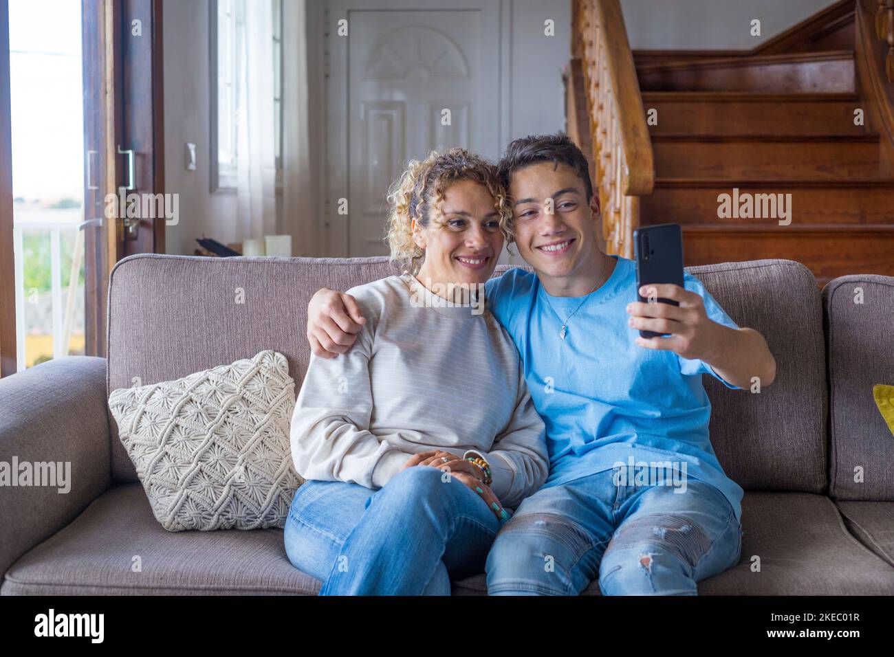 Smiling middle-aged 40s mother rest with grown-up son using smartphone together, happy young man enjoy family weekend with mom taking a selfie, have fun at home Stock Photo