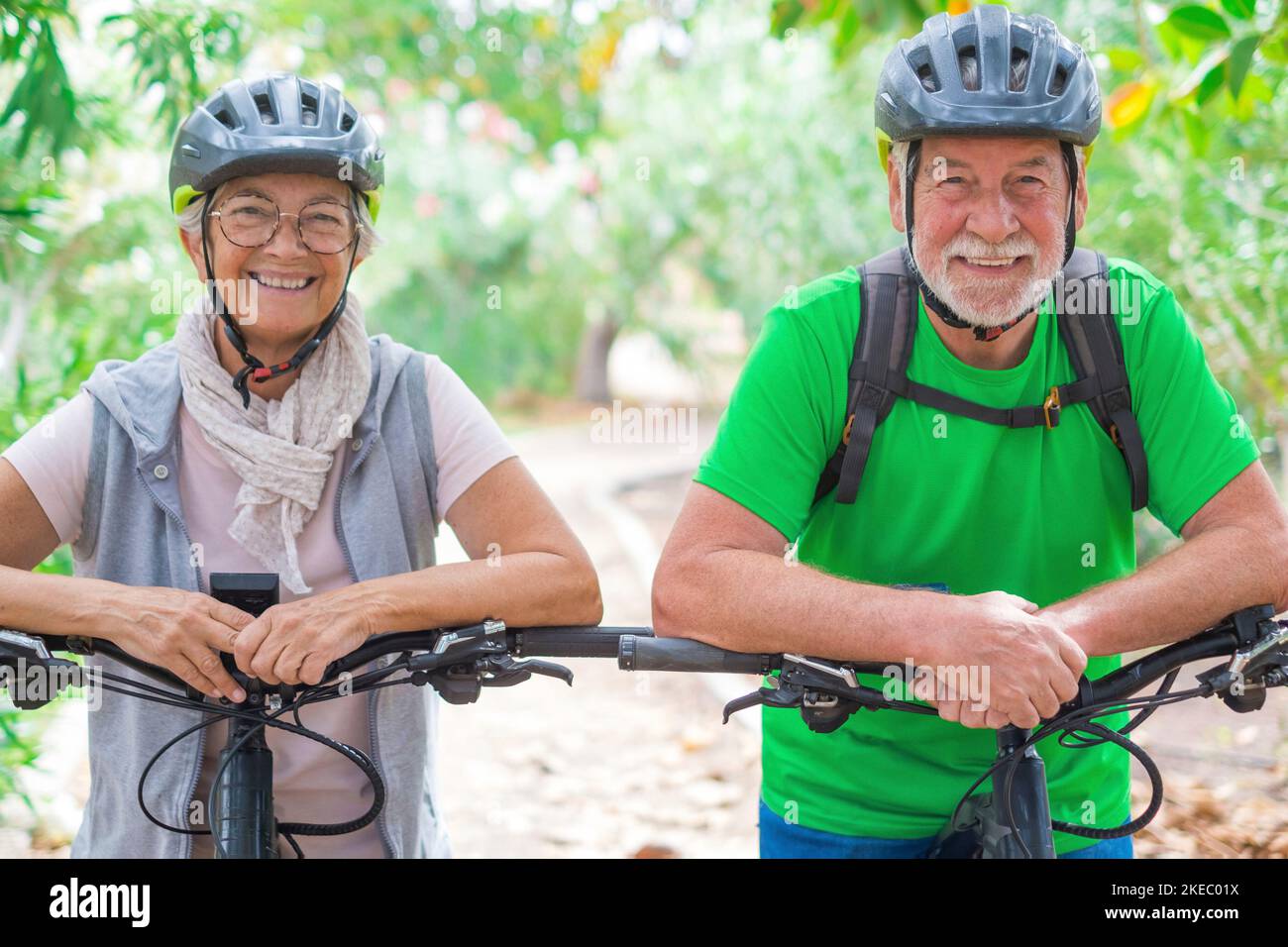Portrait of couple of old and happy in love seniors looking at the camera smiling and having fun with their bikes in the nature outdoors together feeling good and healthy. Stock Photo