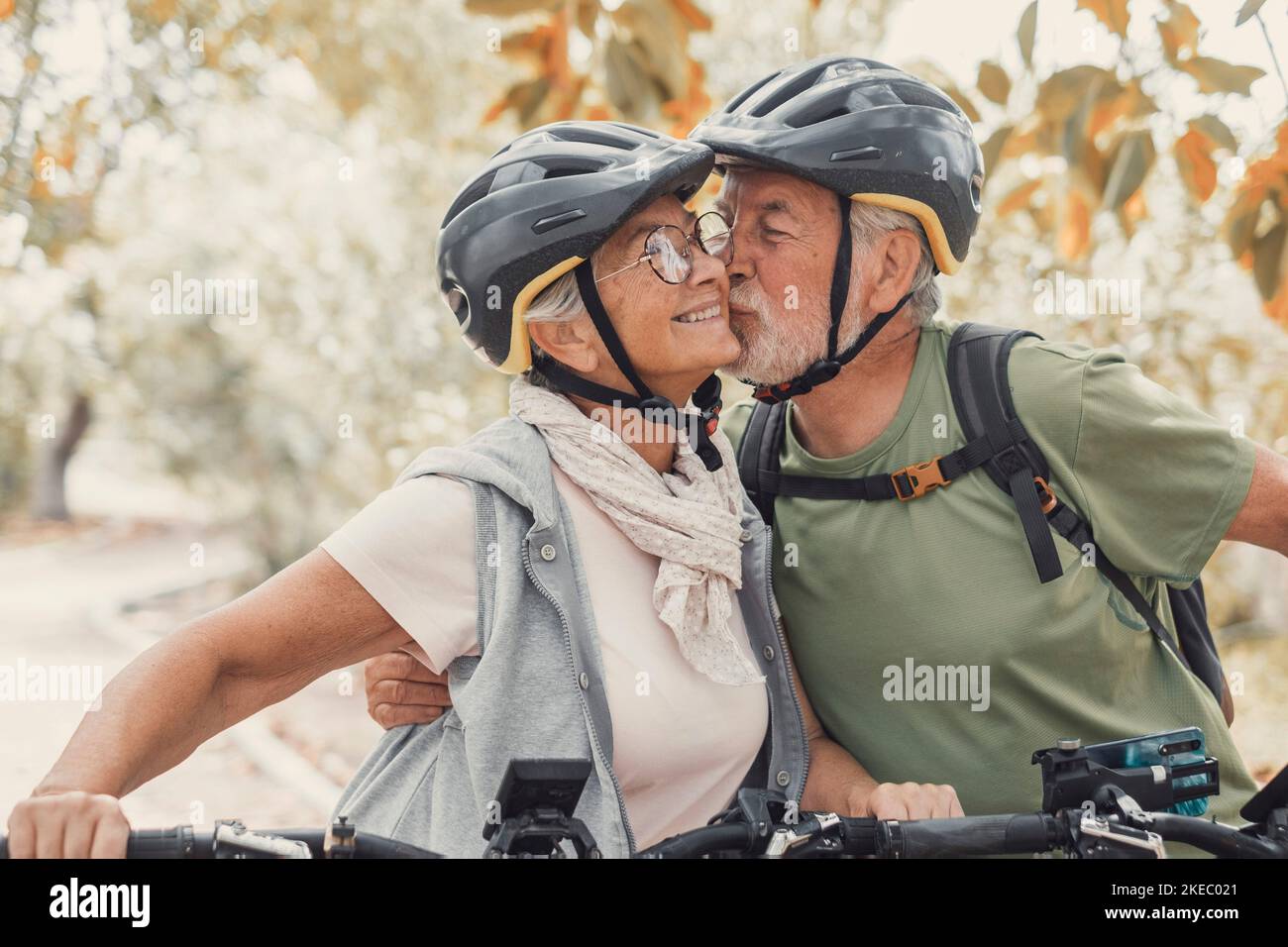 Couple of cute and sweet seniors in love enjoying together nature outdoors having fun with bikes. Old man kissing his wife smiling and feeling good. Stock Photo