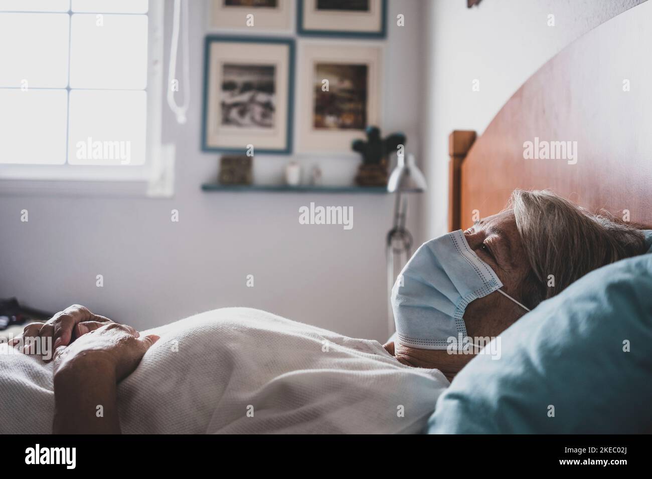 covid-19. one sick mature and retired woman having a bad time in the bed of her home - infected and upset senior wearing mask indoors resting Stock Photo