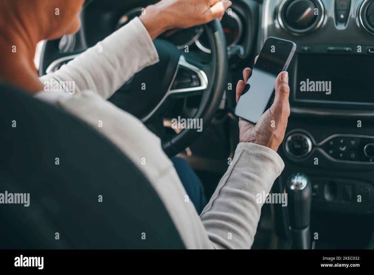 Portrait of one old woman using phone in car while driving in the road risk causing an accident. Close up of hands holding smartphone surfing online texting and chatting. Stock Photo