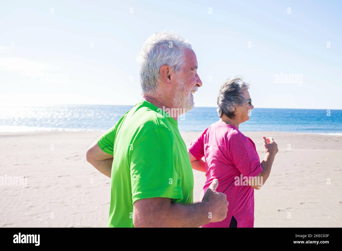 couple of seniors running together at the beach with the sea at the background - two mature people enjoying jogging and smiling Stock Photo