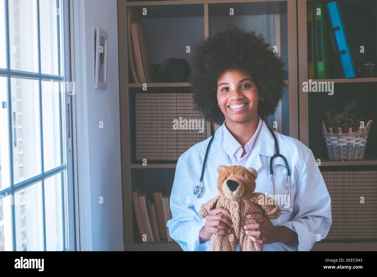 Portrait of young african american female nurse in medical uniform holding stuffed toy showing care love to little patient, happy woman doctor pediatrician posing with teddy bear in hospital or clinic Stock Photo
