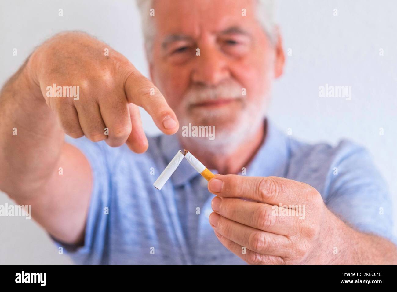 close up of mature man holding a cigar in his hands showing he can leave from the world of the smoke - stop smoking and the smoke kill concept and portait - senior with white wall at the background Stock Photo