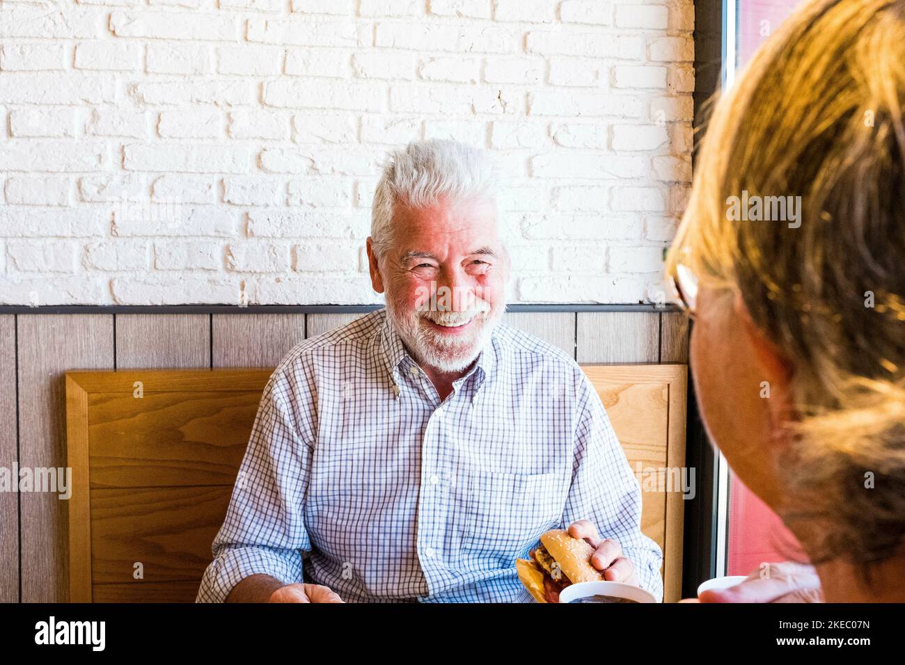 couple of two seniors eating and drinking in a restaurant of fast food together - mature man holding an hamburger and prepared to eat it looking at his wife Stock Photo