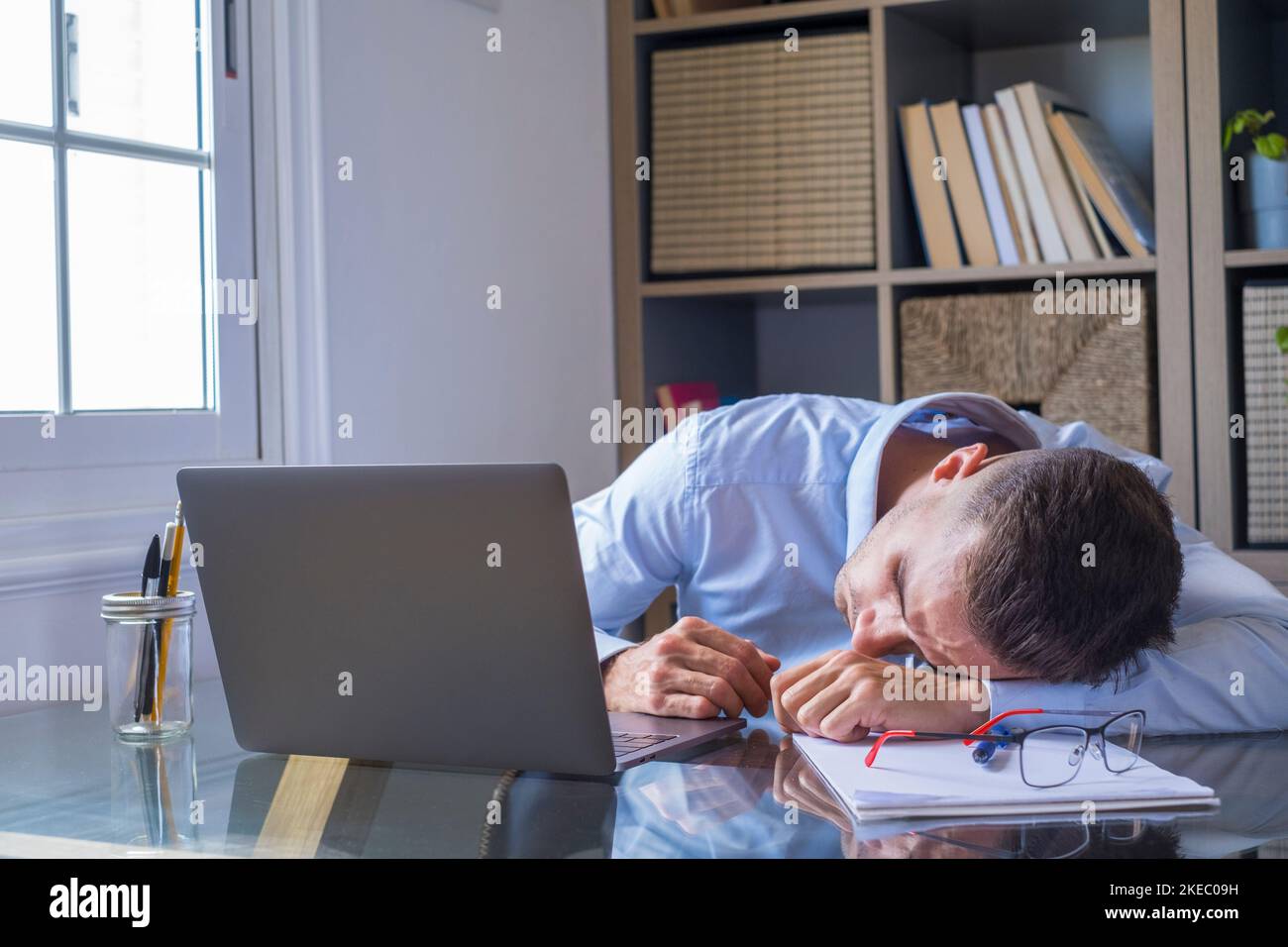 Tired businessman is sleeping at his work desk with laptop computer. Exhausted caucasian guy lying on table with eyes closed falling asleep. Male executive sleeping at workplace, boring routine work Stock Photo