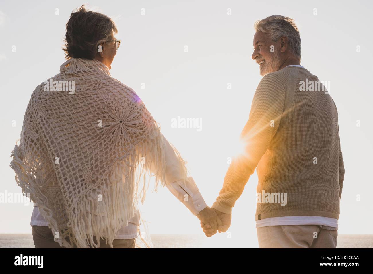 couple of two happy and active seniors having fun and enjoying together summer at the beach walking holding their hands with the sunset at the background Stock Photo