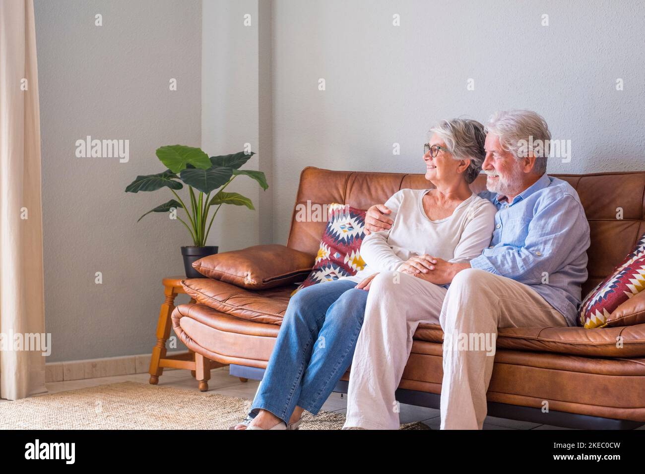 Senior couple relaxing on sofa at home and looking out through window on a bright sunny day. Happy old husband and wife looking away and admiring view sitting on couch at modern house Stock Photo