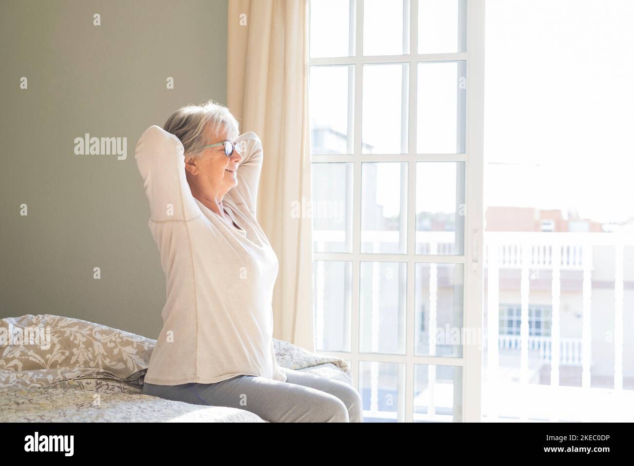 Happy senior woman sitting on bed with hands behind head and eyes closed in bedroom next to window at her apartment. Satisfied old woman stretching hands on bed at home Stock Photo