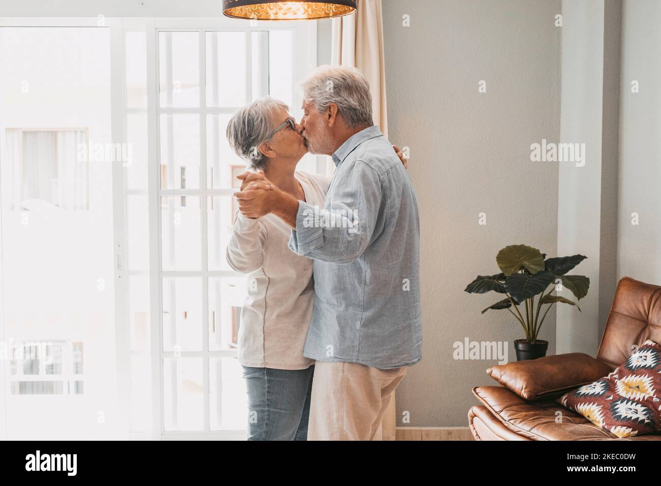 Romantic loving senior couple holding hands enjoying dancing together while kissing each other in the living room of house, couple kissing and embracing while holding hands together at home Stock Photo