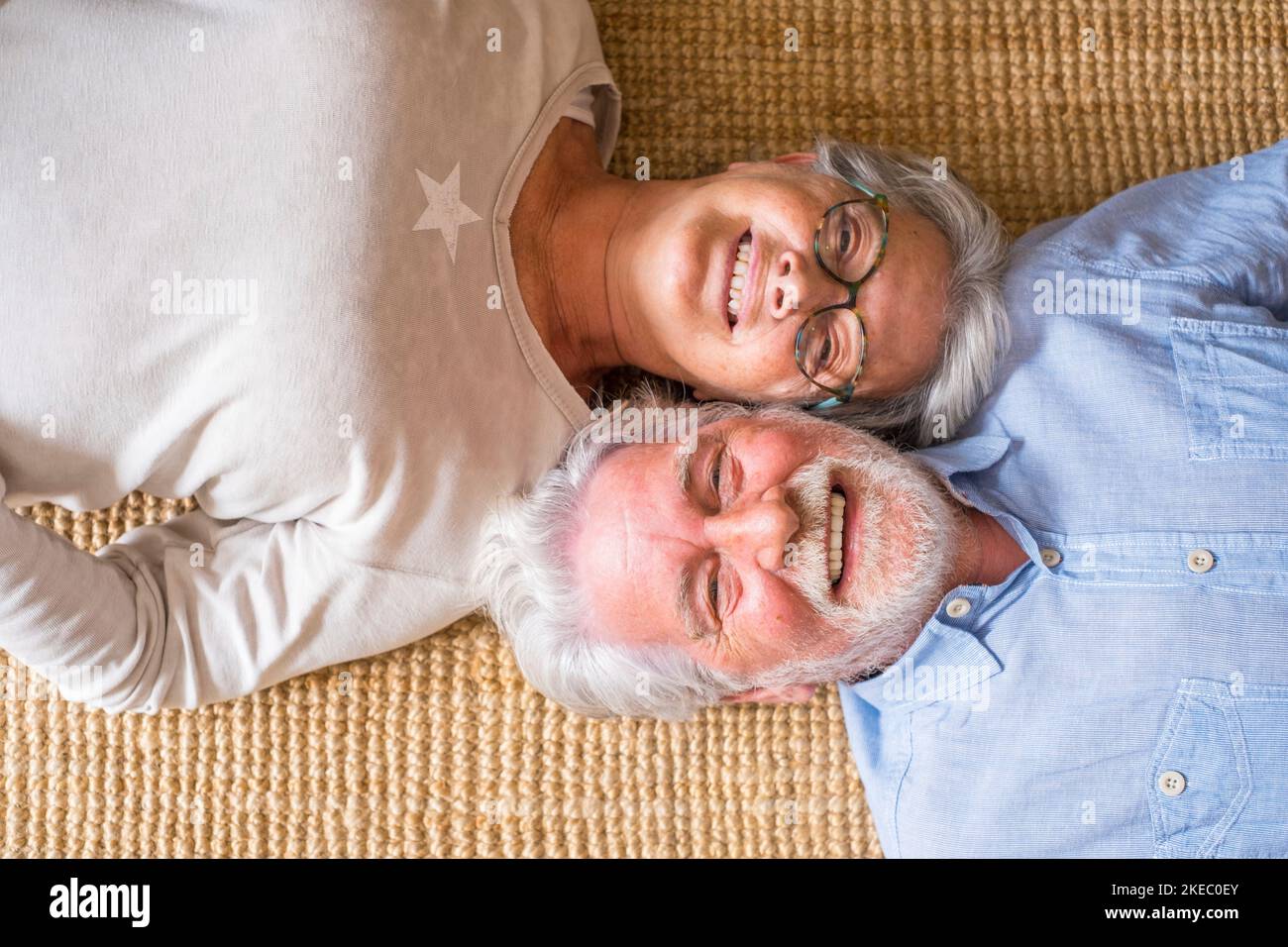 Elderly senior couple smiling while lying on carpet in living room at home, couple relaxing on rug. Top view of happy couple lying down on carpet at home Stock Photo