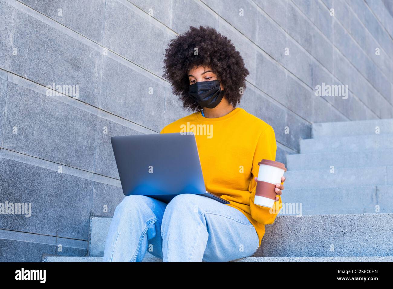 Beautiful African American woman in yellow t-shirt, face mask and curly hair sits on stairs with laptop and coffee mug Stock Photo