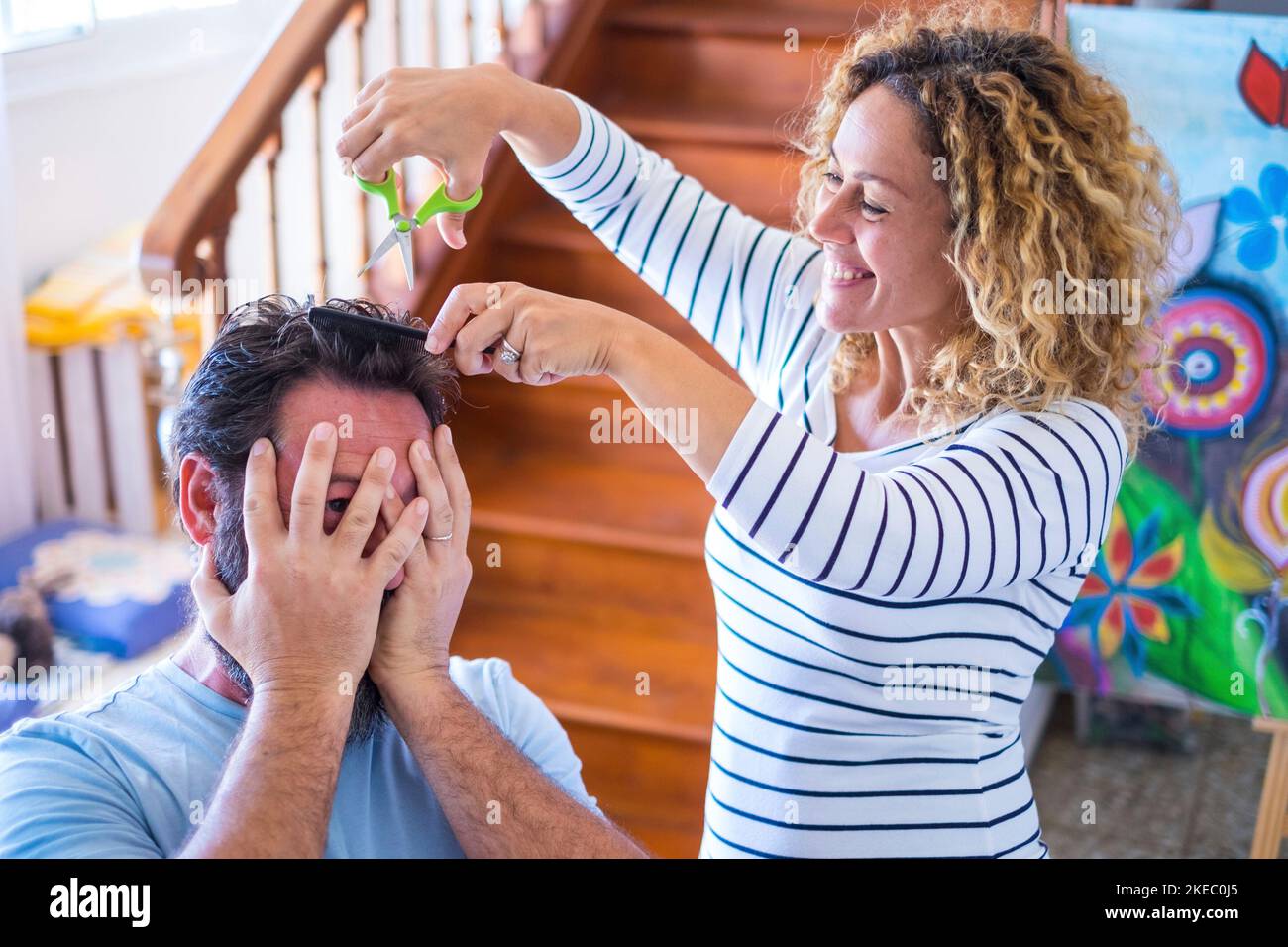 couple of people having fun together at home enjoying and playing with scissors cutting the hair of her husband - happy one curly woman cutting the hairstyle of man Stock Photo