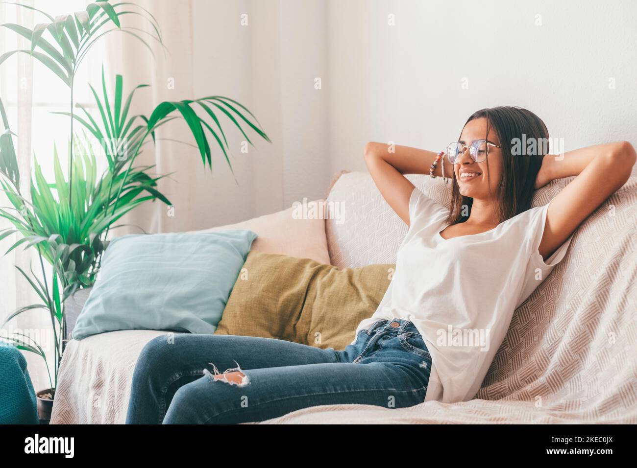 Happy young woman in eyeglasses relaxing on sofa with hands behind head in the living room of her house. Carefree and satisfied lady with torn jeans relaxing on couch at modern apartment Stock Photo