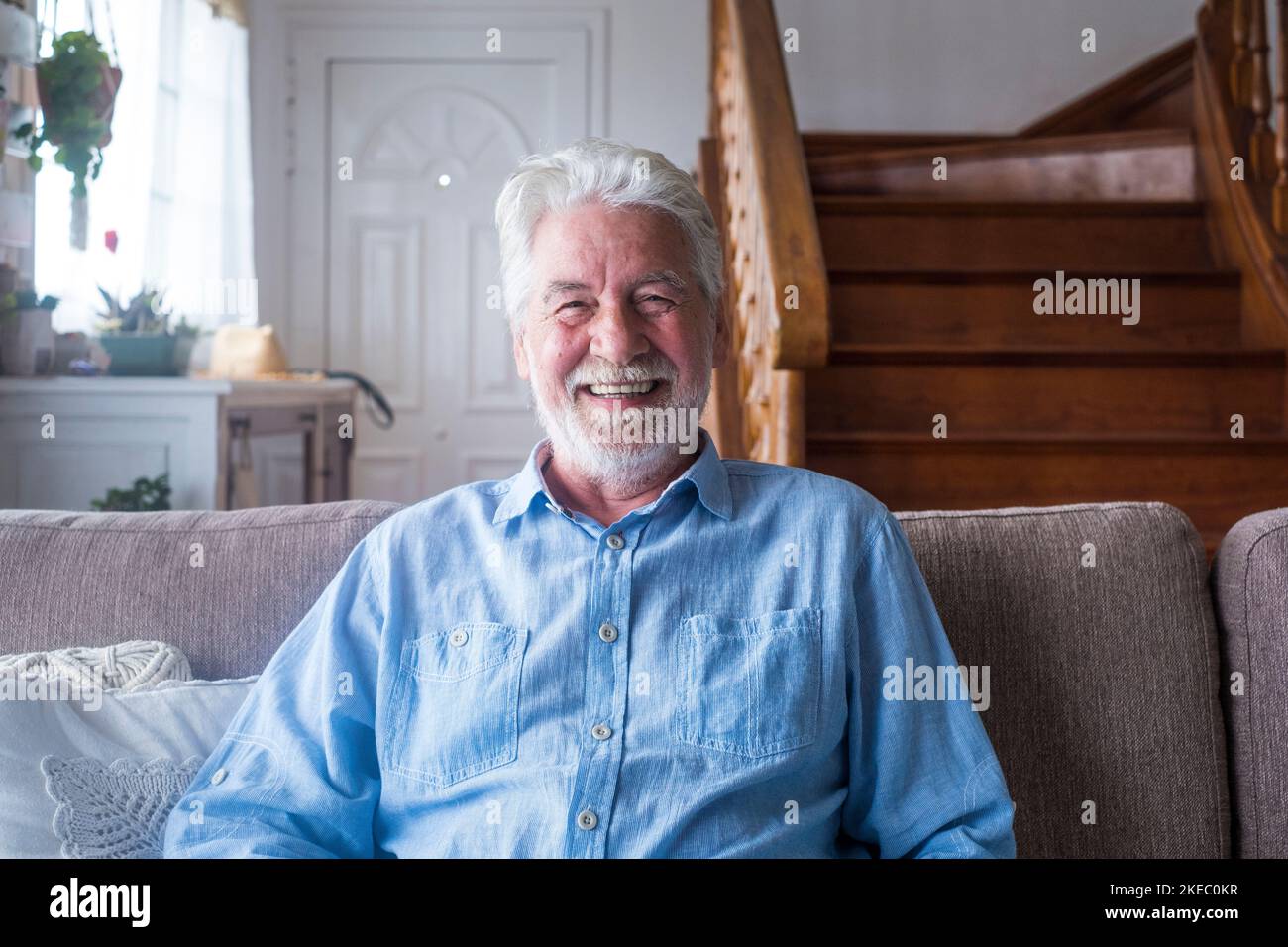 Portrait of happy senior man sitting on sofa in the living room of house in front of wooden staircase. Old male smiling while relaxing on couch and looking at camera Stock Photo