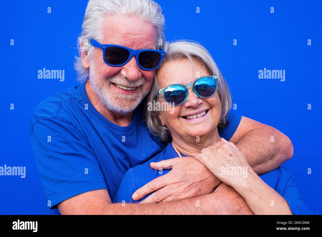 portrait of couple of mature people or seniors together having fun smiling and looking at the camera - woman and man pensioners wearing blue shirt and sunglasses with blue background - trendy colorful and happy people Stock Photo