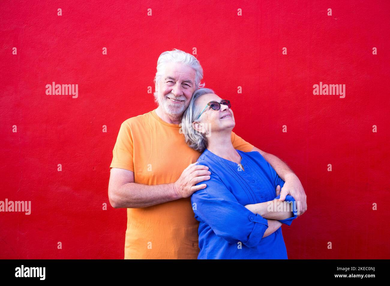 couple of seniors having fun together and smiling - mature man looking at the camera and hugging his wife - red background - portrait sand close up Stock Photo