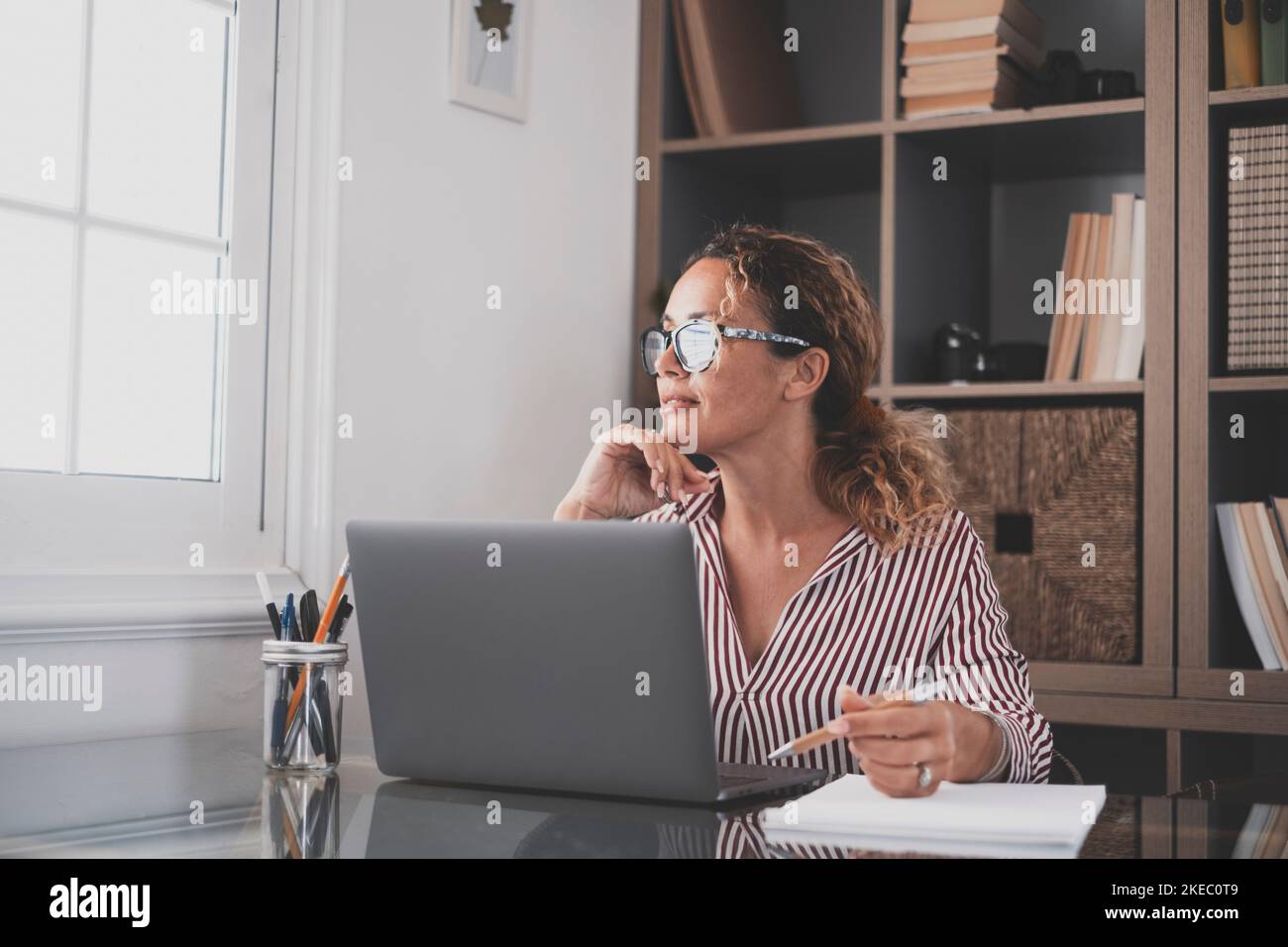 Female business executive lost in thoughts in search for inspiration and looking for new creative ideas. Deramy contemplative caucasian businesswoman looking away while sitting at office desk with laptop Stock Photo
