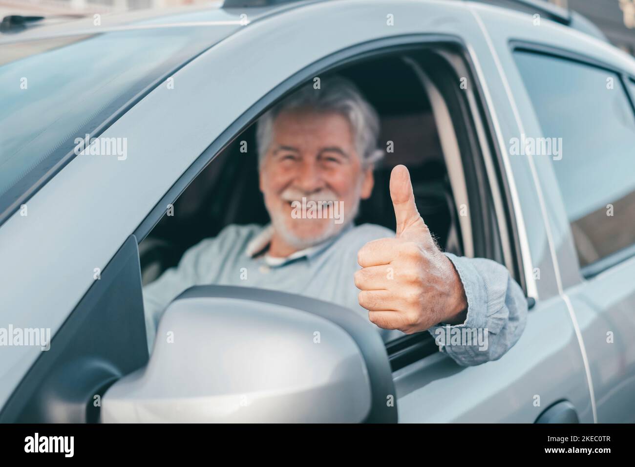 Happy owner looking at the camera with happy face and thumbs up. Handsome bearded mature man sitting relaxed in his newly bought car looking out the window smiling joyfully. One old senior driving and having fun. Stock Photo