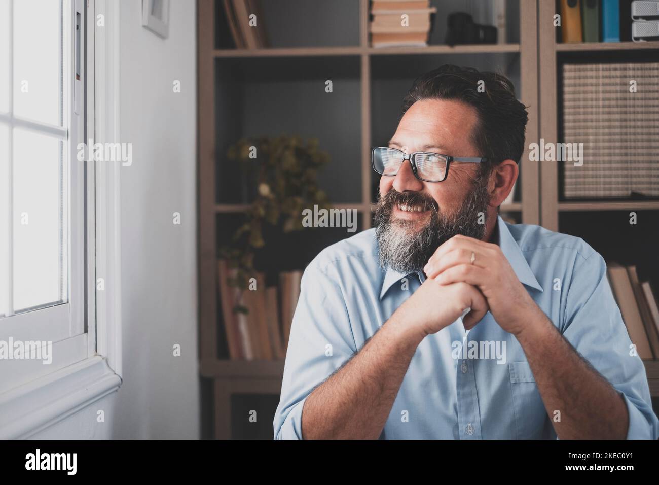 Confident man looking away with big smile. Happy handsome guy looking out through window thinking about the future. Smiling male daydreaming and admiring view from his apartment Stock Photo