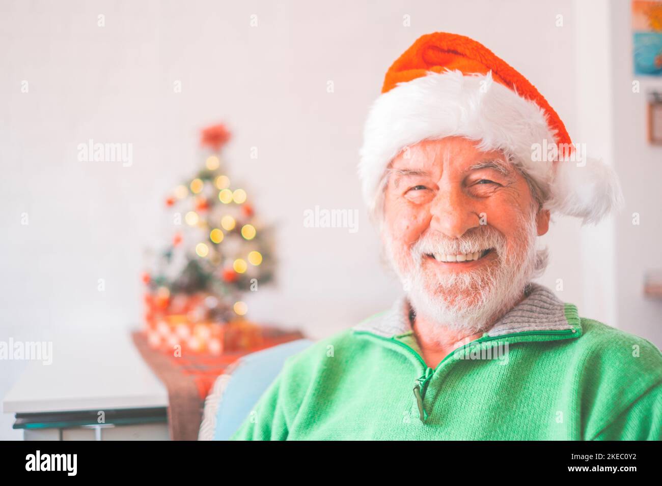 Portrait of smiling retired senior man in santa hat and warm clothing celebrating christmas at home. Old male santa smiling while looking at camera. Elderly man enjoying christmas holiday Stock Photo