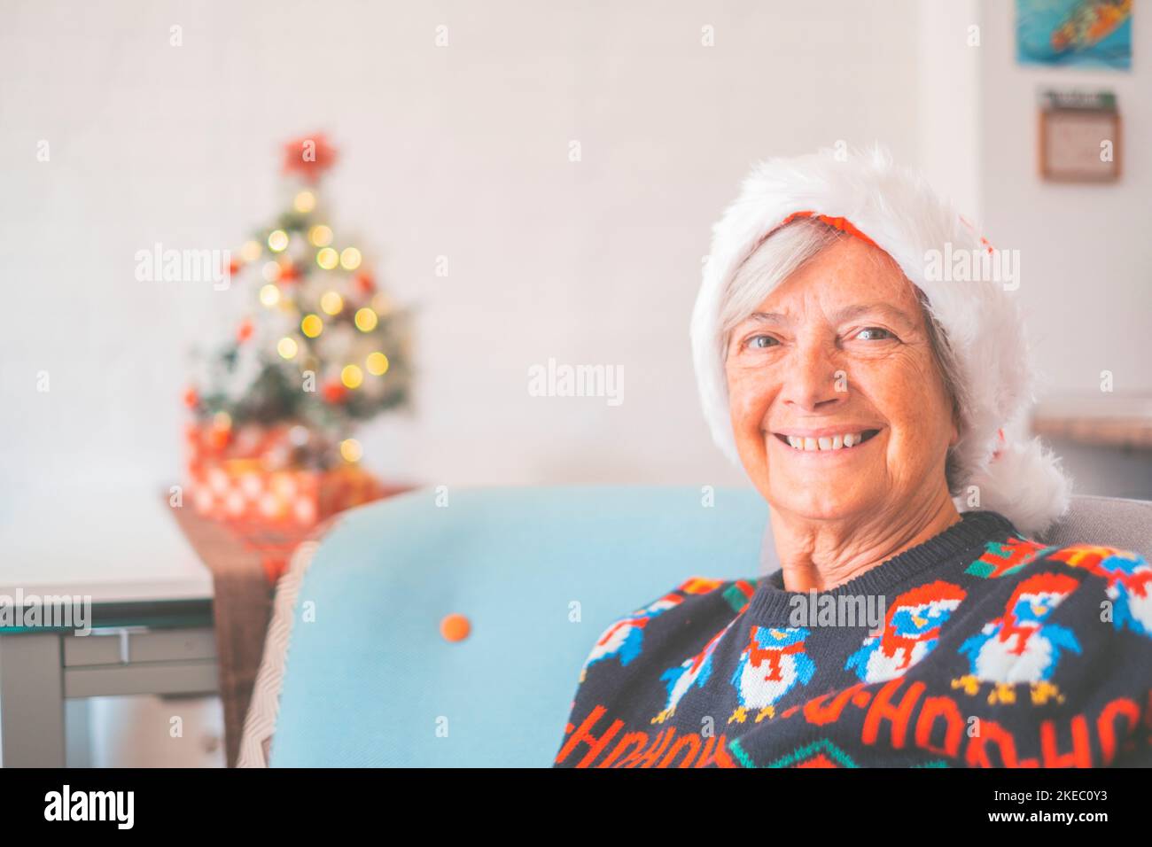 Portrait of smiling retired senior woman in santa hat and warm clothing celebrating christmas at home. Old female santa smiling while looking at camera. Elderly lady enjoying christmas holiday Stock Photo