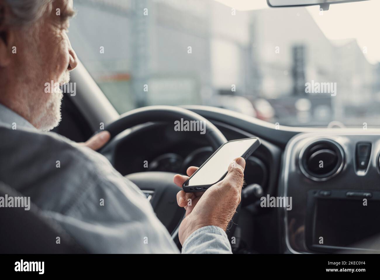 Man looking at mobile phone while driving a car. Old pensioner person distracting texting and chatting. Stock Photo
