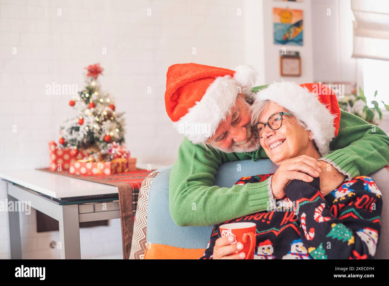 Senior couple in warm clothing and santa hat holding each others hands in front of decorated christmas tree at home. Loving old romantic heterosexual couple celebrating christmas festival together Stock Photo