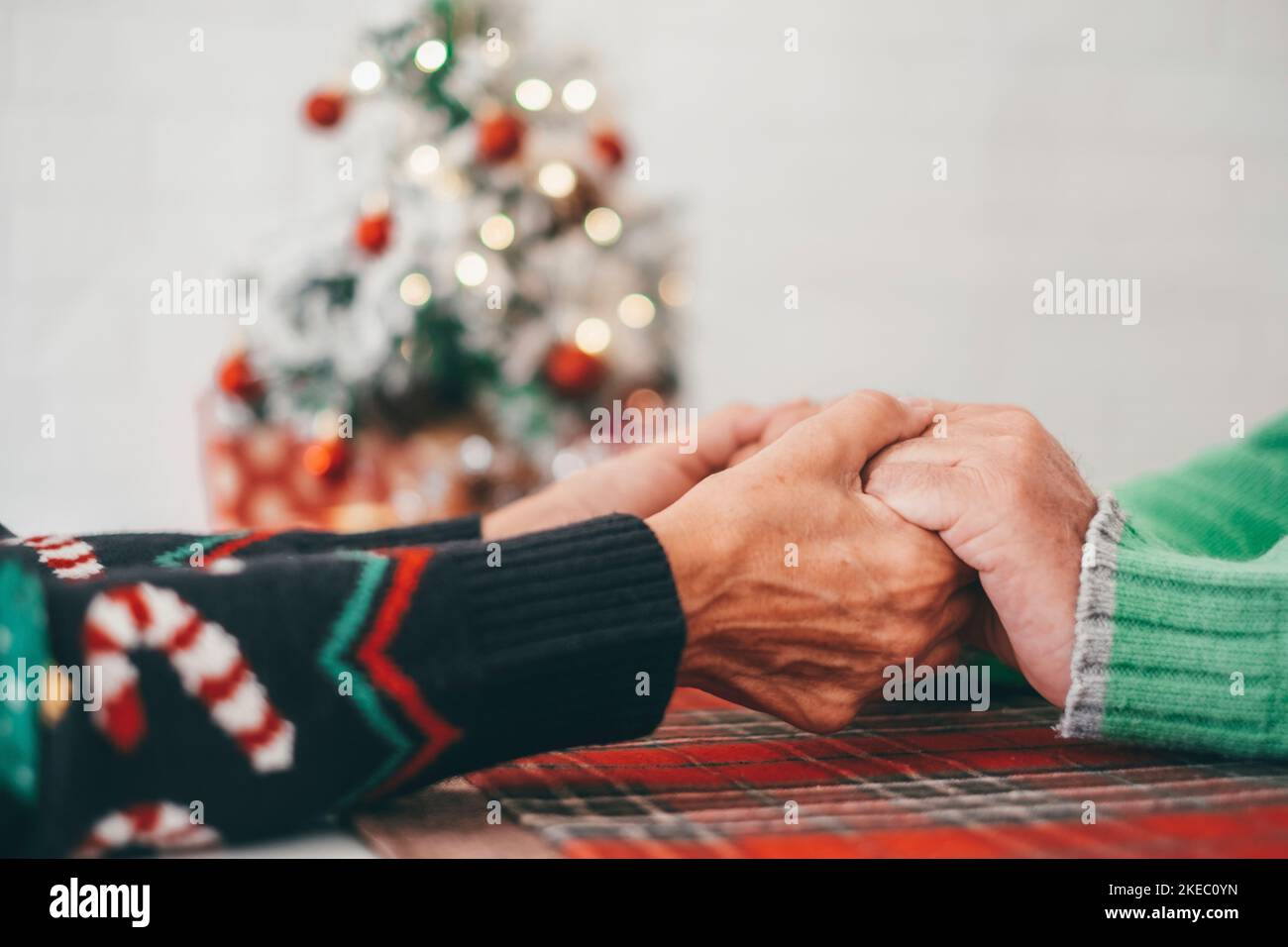 Senior couple in warm clothing holding each others hands in front of decorated christmas tree at home. Loving old romantic heterosexual couple celebrating christmas festival together Stock Photo