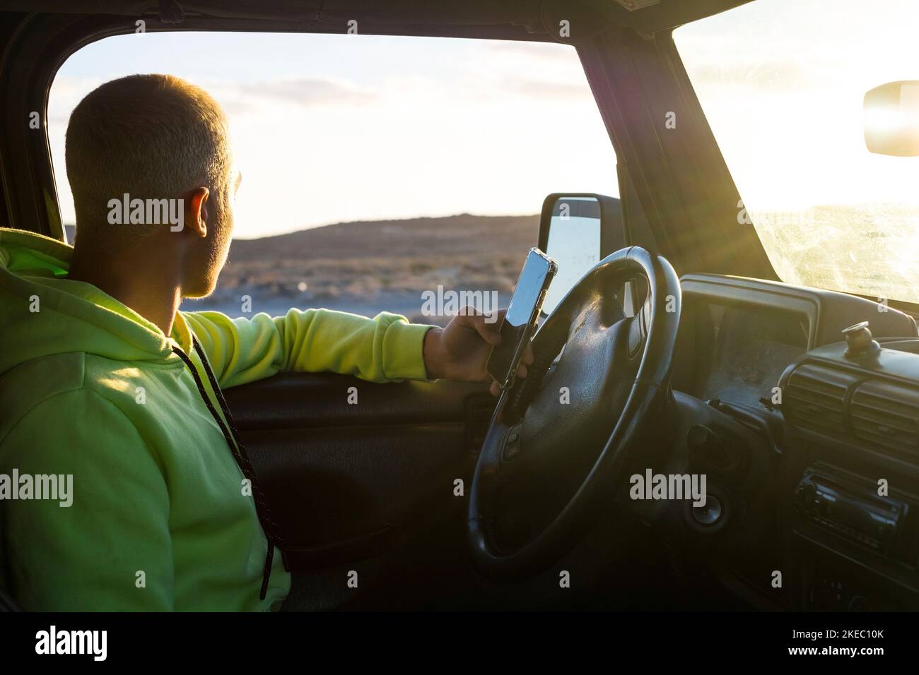 one young man driving his car alone and looking at the amazing sunset at the background while using his phone to searching some maps or social media Stock Photo