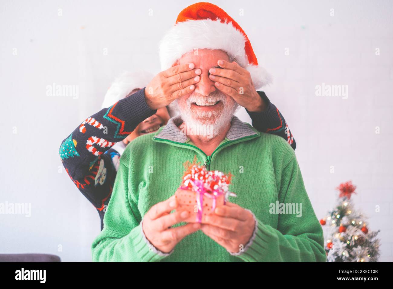 Senior couple in warm clothing and santa hat, woman covering eyes of old husband with a surprise gift for him. Loving old romantic heterosexual couple celebrating christmas festival together Stock Photo