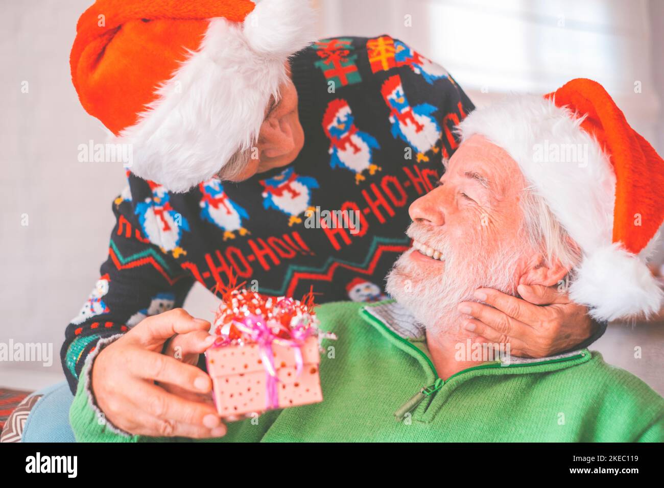 Senior couple in warm clothing and santa hat, woman giving a surprise christmas gift to her husband. Loving old romantic heterosexual couple celebrating christmas festival together Stock Photo