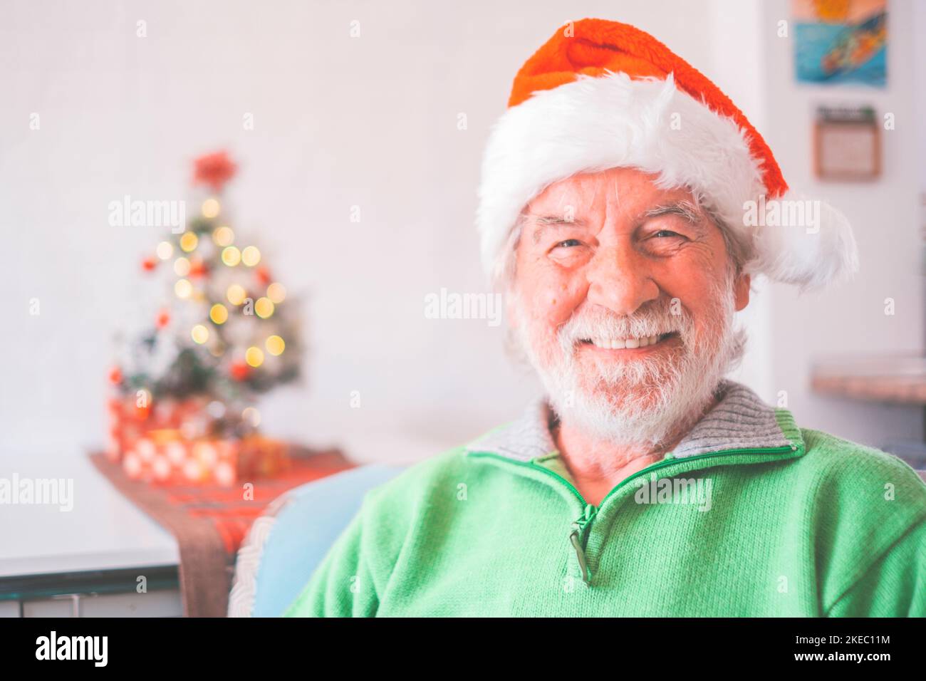 Portrait of smiling retired senior man in santa hat and warm clothing celebrating christmas at home. Old male santa smiling while looking at camera. Elderly man enjoying christmas holiday Stock Photo