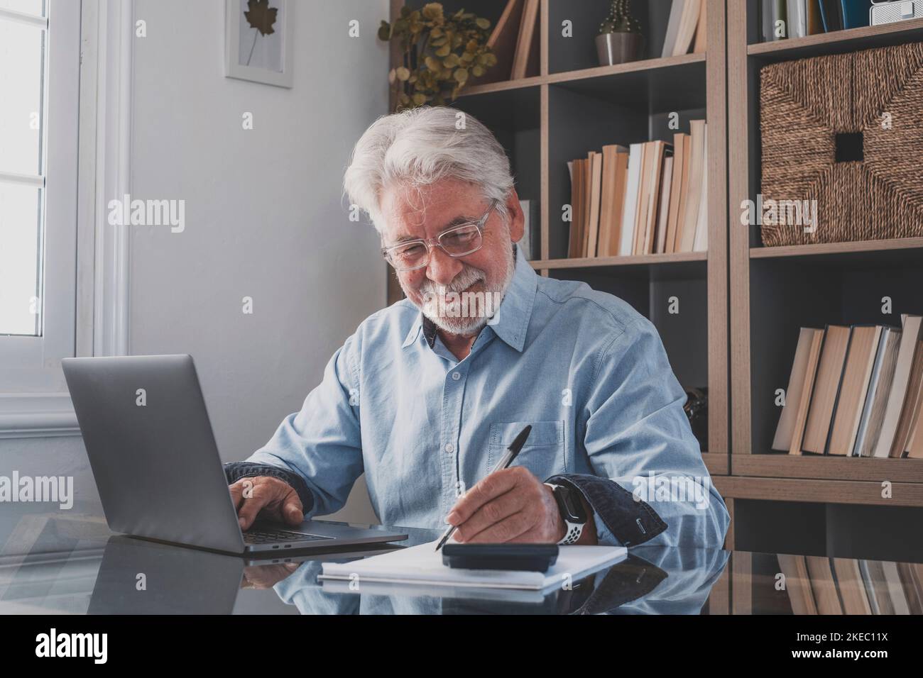 Happy old caucasian businessman smiling working online watching webinar podcast on laptop and learning education course conference calling make notes sit at work desk, elearning concept Stock Photo