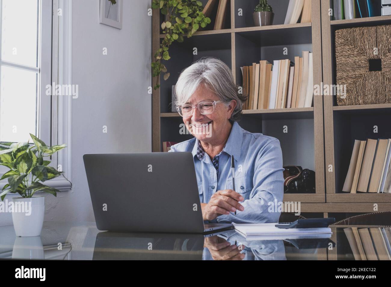 Happy old caucasian businesswoman smiling working online watching webinar podcast on laptop and learning education course conference calling make notes sit at work desk, elearning concept Stock Photo