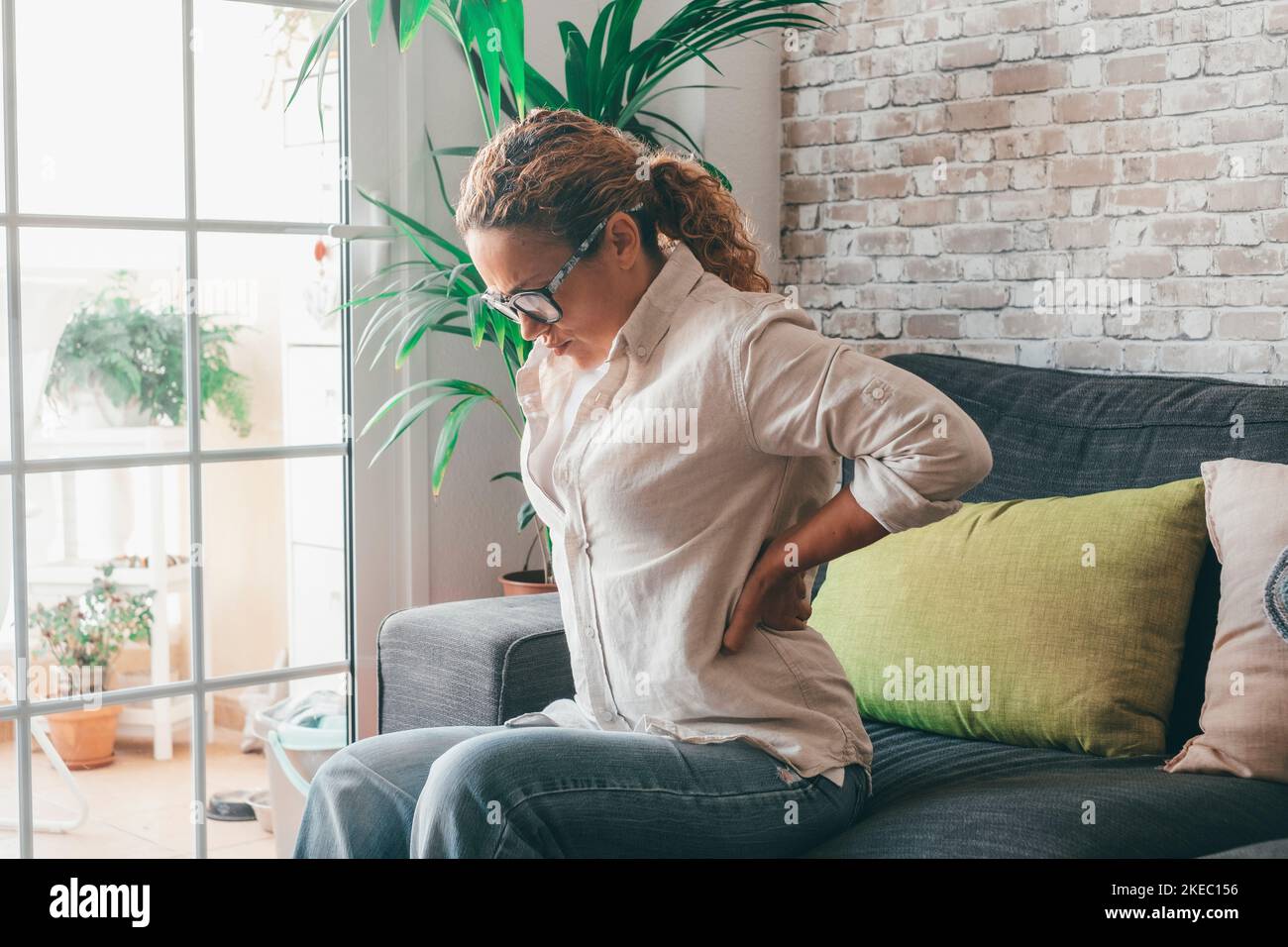 Tired young woman suffering from severe backache sitting on sofa in the living room of house, Caucasian female sitting on couch touching lower back feeling discomfort at home Stock Photo