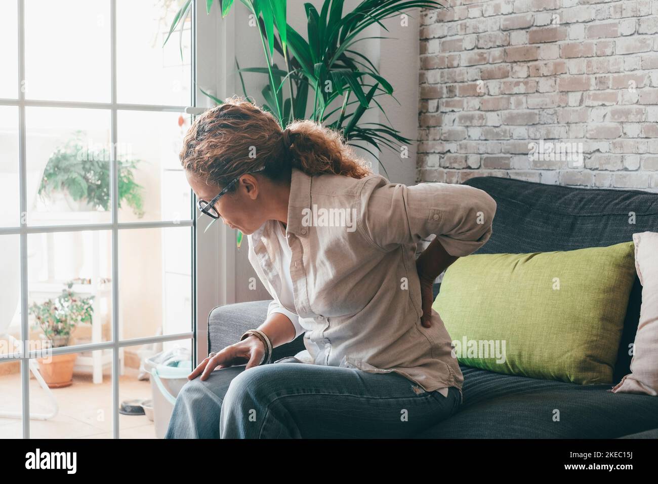 Unhealthy curly woman sit on couch at home stretching suffering from back problems, unwell overworked female touch massage lower spine, having muscular pain or strain. Incorrect posture concept Stock Photo