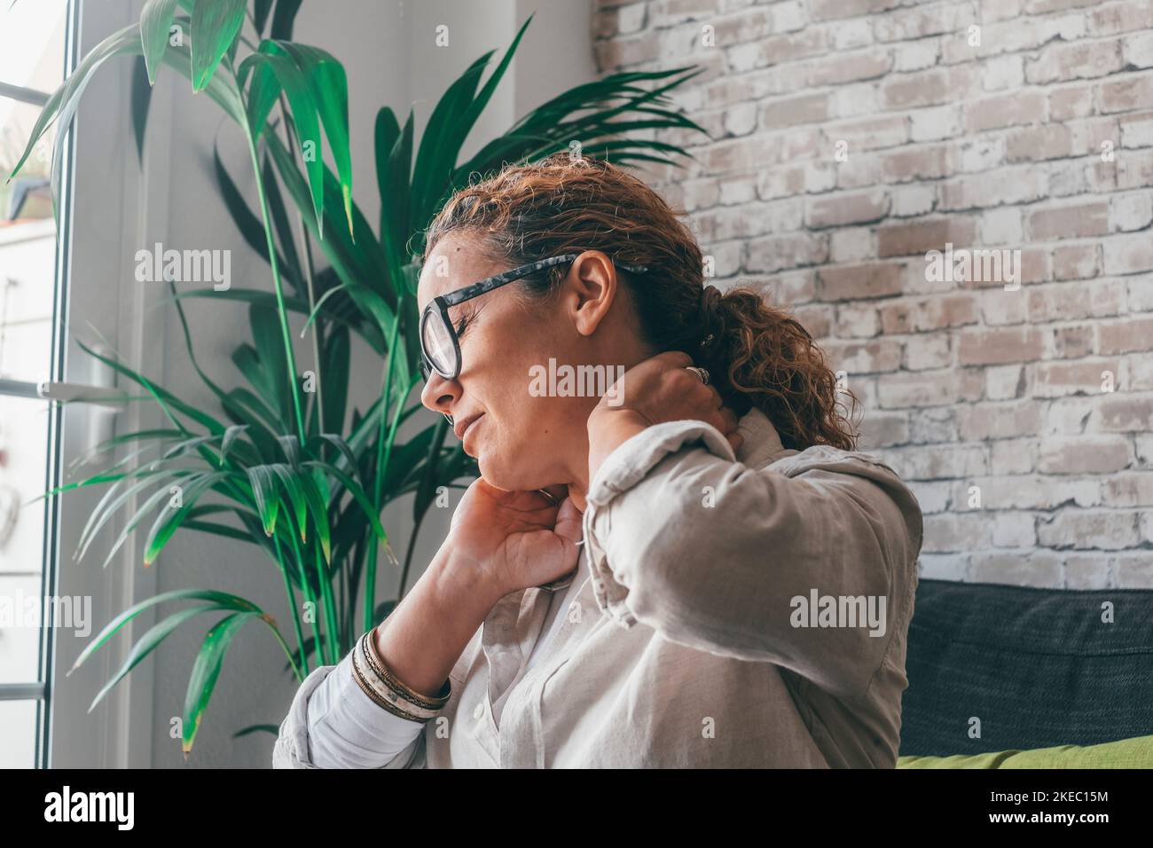 Exhausted young woman suffering from strong neck ache while sitting on sofa in the living room of house, Caucasian female sitting on couch touching and massaging tensed neck muscles feeling discomfort at home Stock Photo