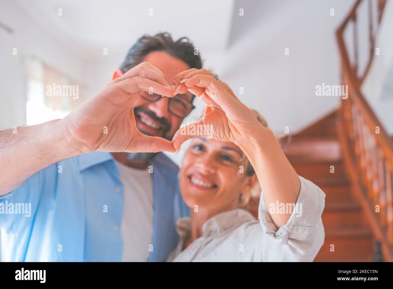 Happy caucasian couple embracing each other in the living room and making heart shape symbol with hands. Loving husband and wife gesturing love sign with hands. Affectionate man and woman spending leisure time together at home Stock Photo