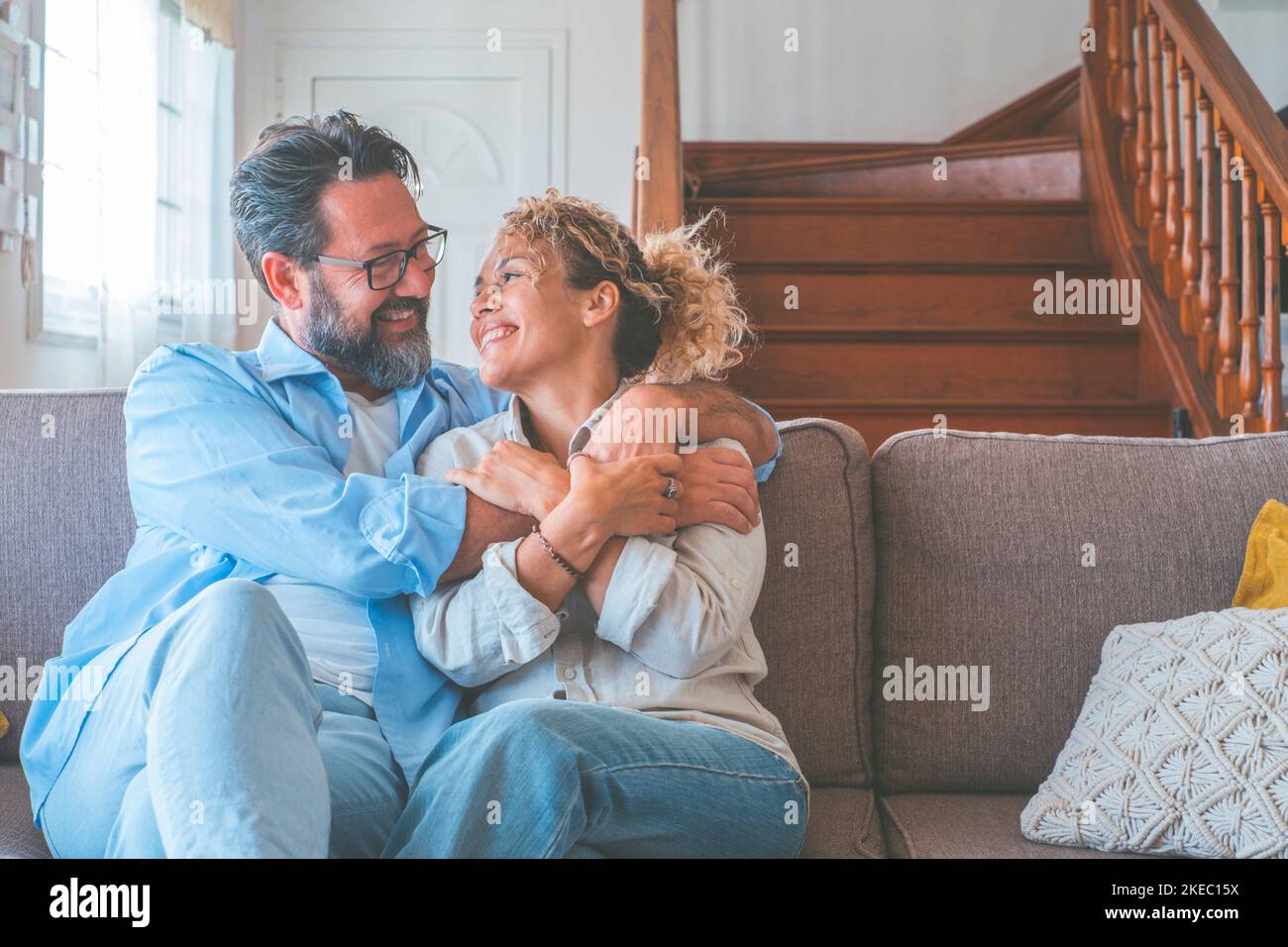 Happy caucasian couple looking at each other sitting on sofa in the living room. Loving husband and wife embracing each other sitting on couch at home. Man and woman spending leisure time at modern apartment Stock Photo